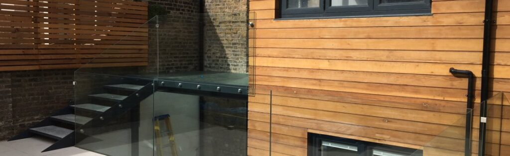 KP Glazing London Modern outdoor patio with wooden panelling, glass railings, black metal stairs, and a brick wall. A window and black drainpipe are visible on the wooden wall. A yellow step ladder is beneath the glass-walled area.