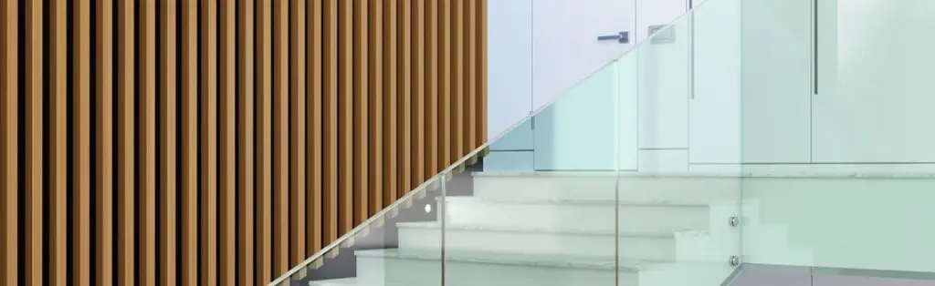 KP Glazing London A modern staircase with white steps and a glass balustrade is next to a wall featuring vertical wooden slats in a contemporary interior space.