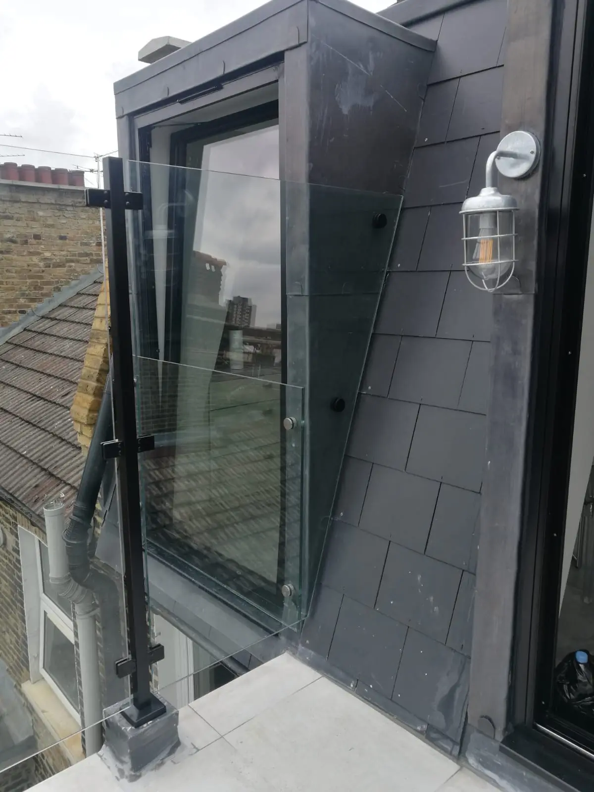 KP Glazing London A modern rooftop balcony with a black-framed glass door, grey slate tiles, a glass balustrade, and a wall-mounted outdoor light. Nearby are brick buildings and a cloudy sky in the background.