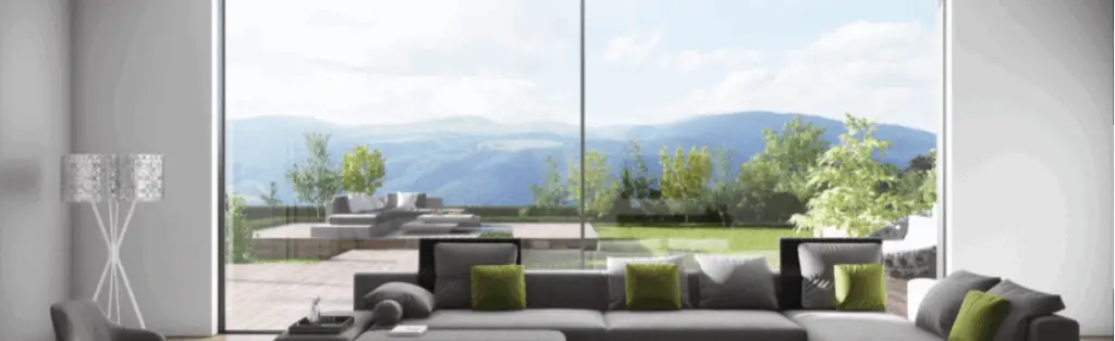 KP Glazing London Modern living room with a large grey sectional sofa, green accent cushions, and floor-to-ceiling windows offering a scenic view of a patio, lush greenery, and distant mountains.