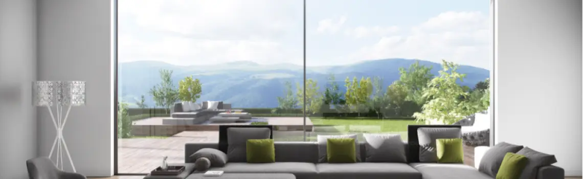 KP Glazing London Modern living room with a large grey sectional sofa, green accent cushions, and floor-to-ceiling windows offering a scenic view of a patio, lush greenery, and distant mountains.