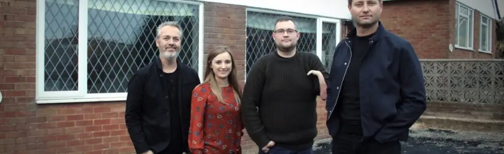 KP Glazing London Four people stand outside in front of a brick building with large windows. Three men and one woman are casually dressed and facing the camera, some with hands in pockets, on a paved area.