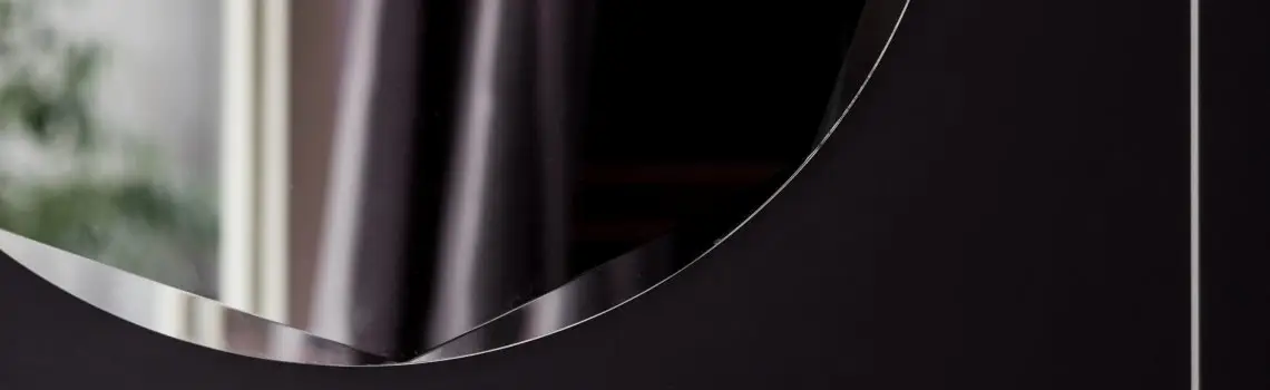 KP Glazing London A close-up of a modern, dark-framed mirror with a curved edge, reflecting part of a window and some greenery in the background.