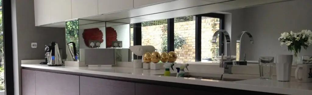 KP Glazing London Modern kitchen with white worktops, dark cupboards, mirrored splashback, and large windows. Various appliances, a vase of white flowers, gold-coloured decor, and cleaning supplies are visible on the counter.