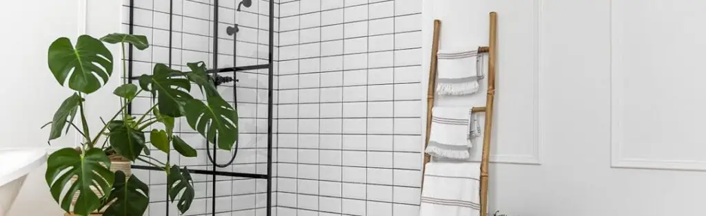 KP Glazing London A modern bathroom with white square tiles, a glass shower enclosure, a leafy green plant, and a wooden ladder holding neatly folded white towels.