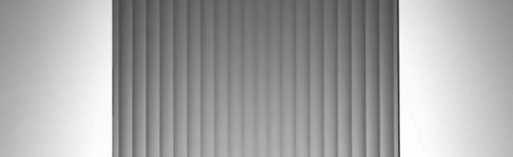 KP Glazing London Vertical grey window blinds partially closed, with light softly illuminating the sides of the wall behind them.