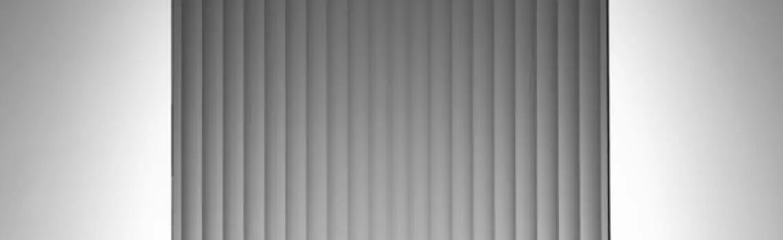 KP Glazing London Vertical grey window blinds partially closed, with light softly illuminating the sides of the wall behind them.