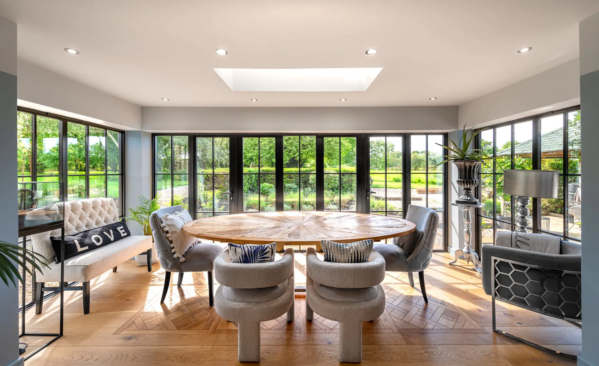 KP Glazing London Bright dining area with large windows overlooking greenery, featuring a round wooden table, upholstered chairs, a cushioned bench with “LOVE” cushions, plants, and parquet wood flooring. Natural light fills the space.