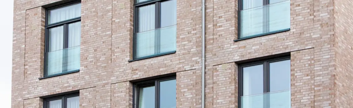 KP Glazing London The image shows a section of a modern brick building façade with large windows, each featuring a glass balcony balustrade. The windows are arranged in a grid pattern across the building.