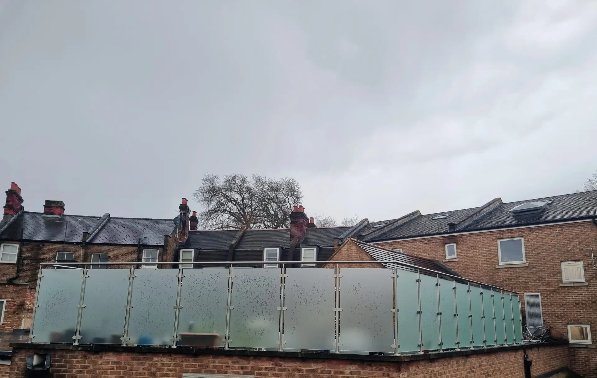 KP Glazing London A row of brick buildings with dark roofs under a cloudy, overcast sky. The closest building has a frosted glass balcony or barrier, and there are leafless trees visible in the background.