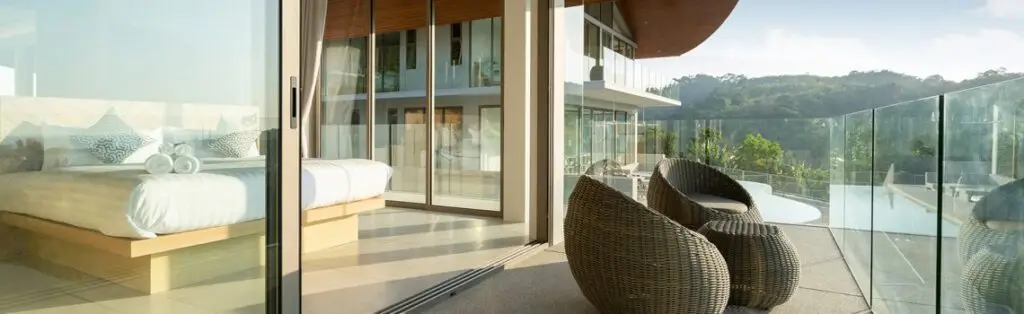KP Glazing London A modern bedroom opens onto a spacious balcony with wicker chairs and a glass balustrade, overlooking greenery and a pool under clear skies.
