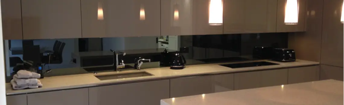 KP Glazing London Modern kitchen with glossy beige cupboards, a light worktop, under-cupboard lighting, a built-in sink, black appliances, and three pendant lights hanging above an island. Towels are stacked on the worktop.
