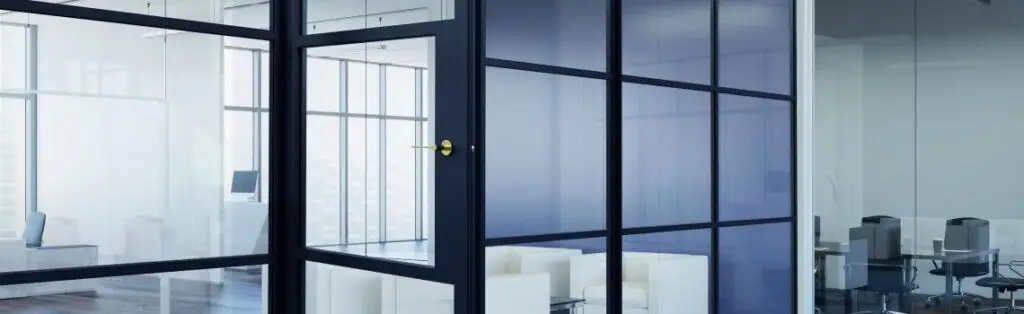 KP Glazing London Modern office space with glass walls and doors, featuring a small meeting room with white chairs inside and an adjacent open workspace with desks and office chairs visible through the transparent partitions.