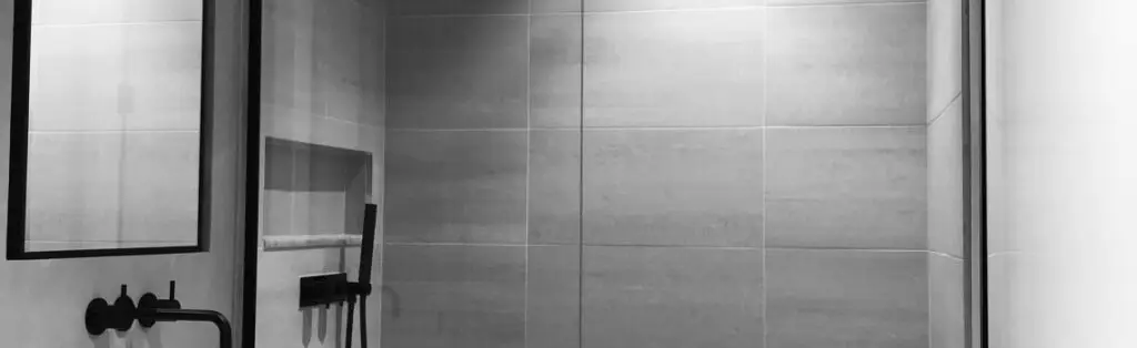 KP Glazing London Black and white photo of a modern bathroom featuring a minimalist shower with a glass door, a large wall mirror with a black frame, a wall niche, and black fittings. The walls are tiled with large rectangular tiles.