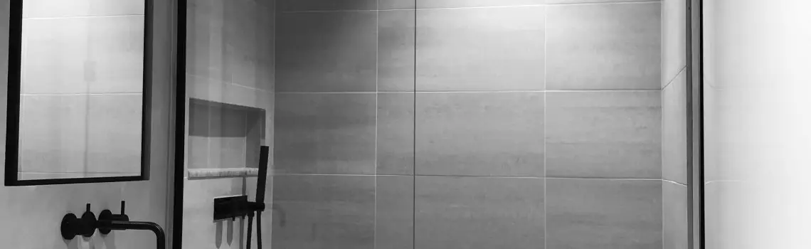 KP Glazing London Black and white photo of a modern bathroom featuring a minimalist shower with a glass door, a large wall mirror with a black frame, a wall niche, and black fittings. The walls are tiled with large rectangular tiles.