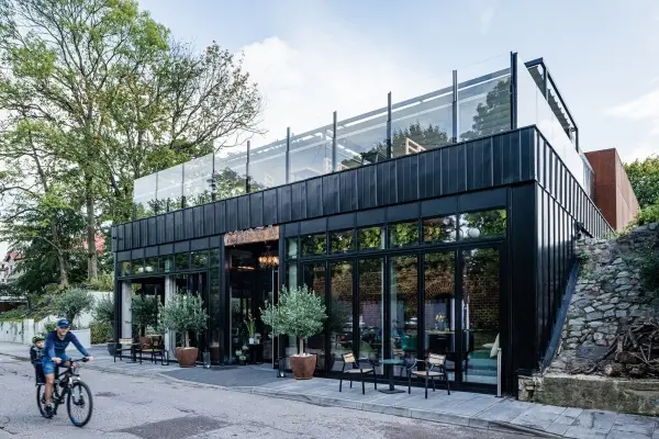 KP Glazing London A modern black building with large glass windows and a rooftop terrace is shown. In front, there are potted plants and outdoor seating. A person wearing a helmet cycles past on the street. Trees and a stone wall are nearby.