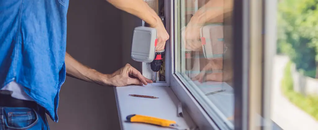 KP Glazing London A person uses a power drill to install or repair a window frame from inside a building. A utility knife and some tools are on the windowsill. Greenery is visible outside the window.