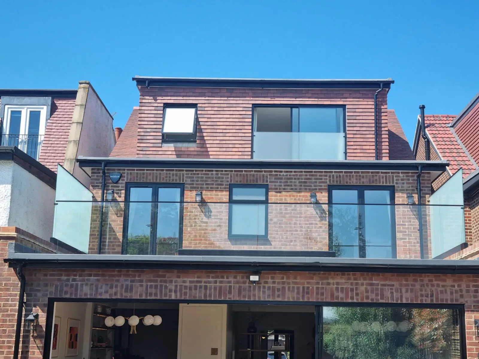 KP Glazing London A modern brick house with large glass doors and windows, a glass balcony, and a loft extension with tiled roof under a clear blue sky. The lower level has an open area leading inside.