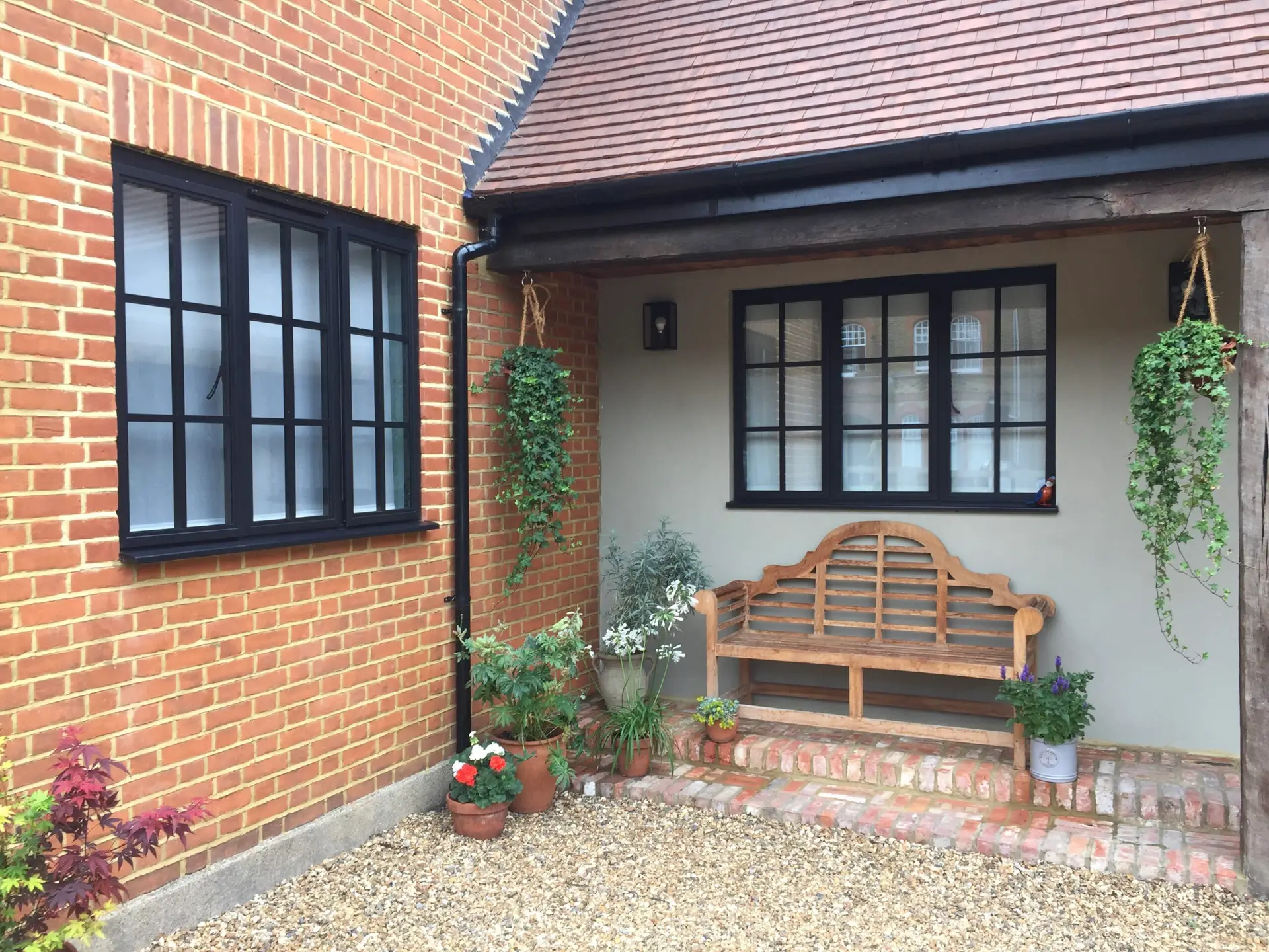 KP Glazing London A wooden bench sits on a brick porch by a house with red brick walls and black-framed windows. Potted plants and hanging baskets with green vines decorate the entrance. Gravel covers the ground.