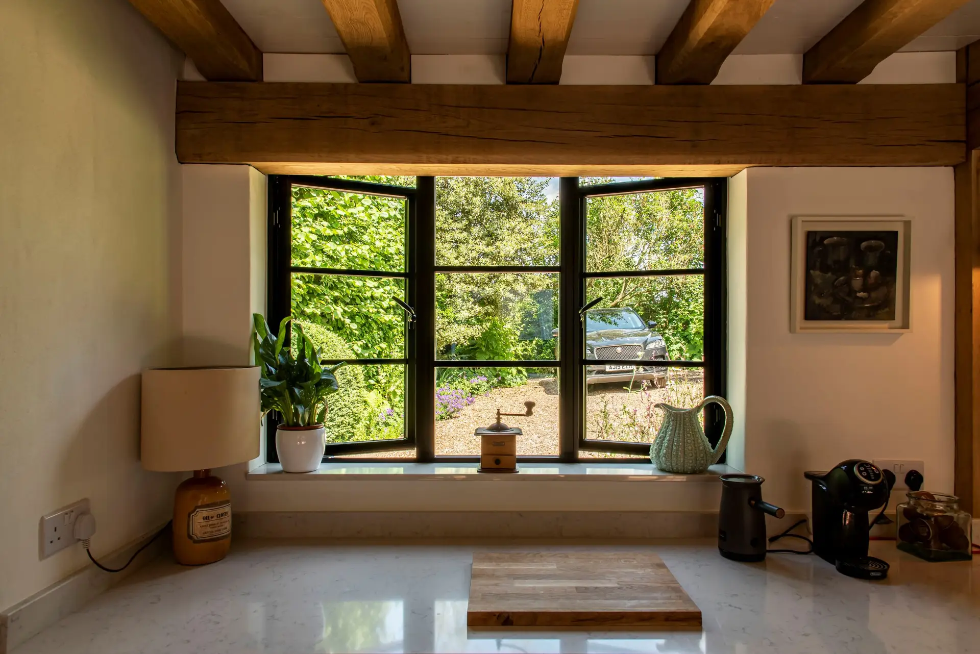 KP Glazing London A kitchen worktop with a lamp, potted plant, coffee maker, kettle, and small canister in front of a wide window overlooking a garden with greenery and a parked car outside.