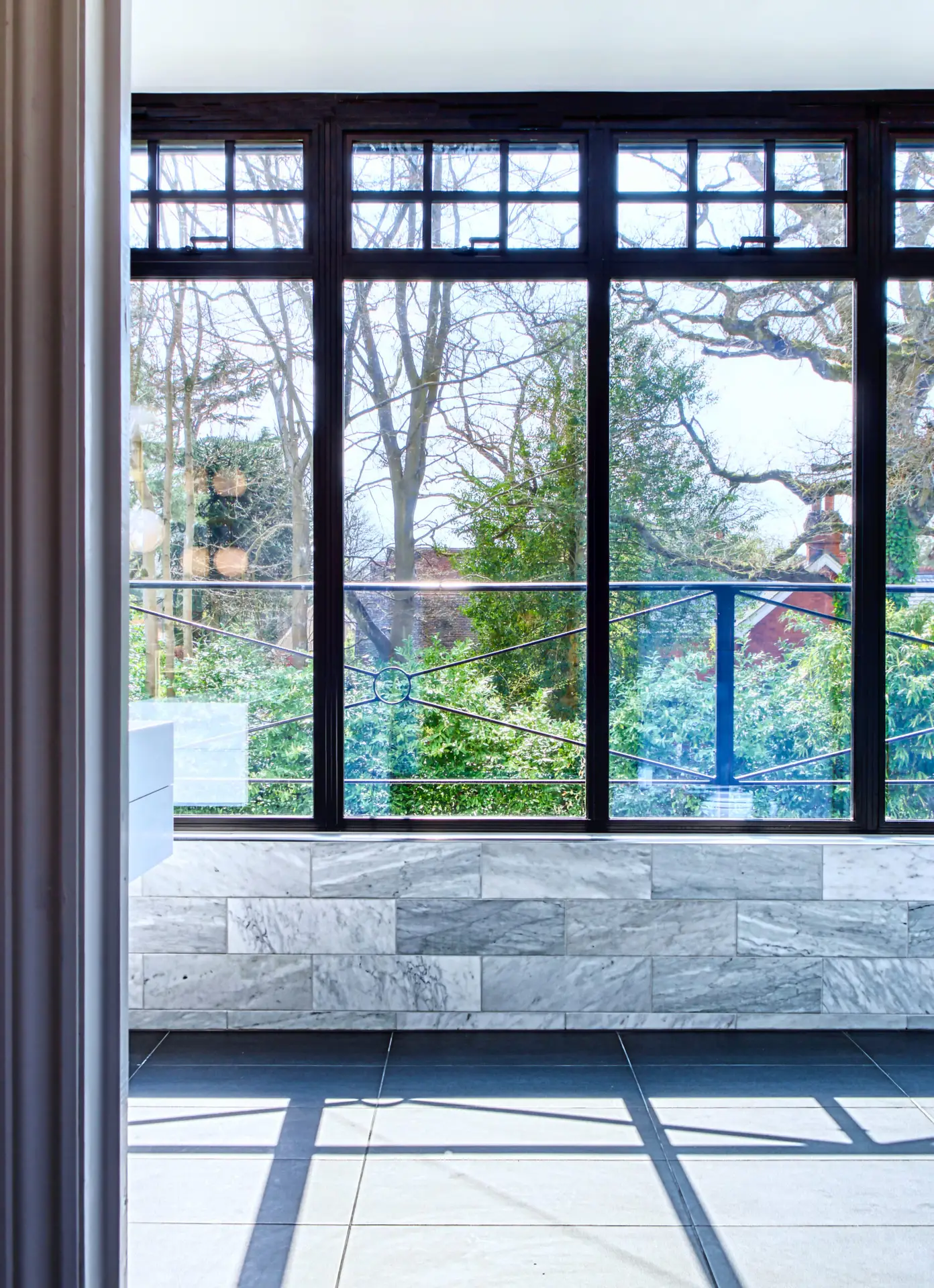 KP Glazing London Large windows with black frames overlook a lush, green garden with trees. Sunlight streams onto grey tiled floors and a half-wall with white marble tiles inside a modern, bright room.