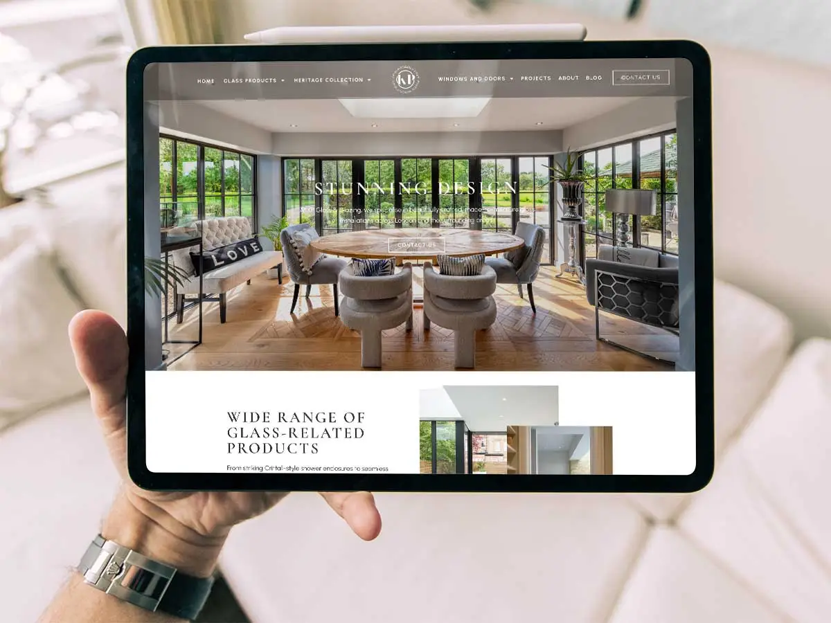 KP Glazing London A hand holding a tablet displaying a modern interior design website featuring a large, stylish dining area with glass walls, elegant chairs, and natural light, with the website navigation menu visible at the top.