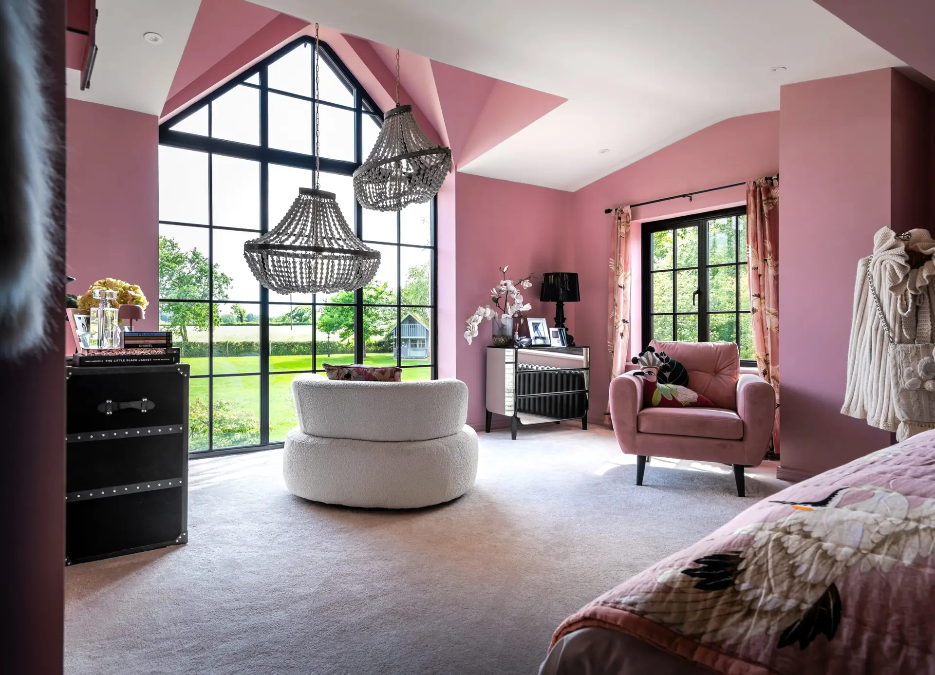 KP Glazing London Bright, modern bedroom with pink walls and ceiling, large black-framed windows, a round white chair, a pink armchair, crystal chandeliers, and a view of a green garden outside.