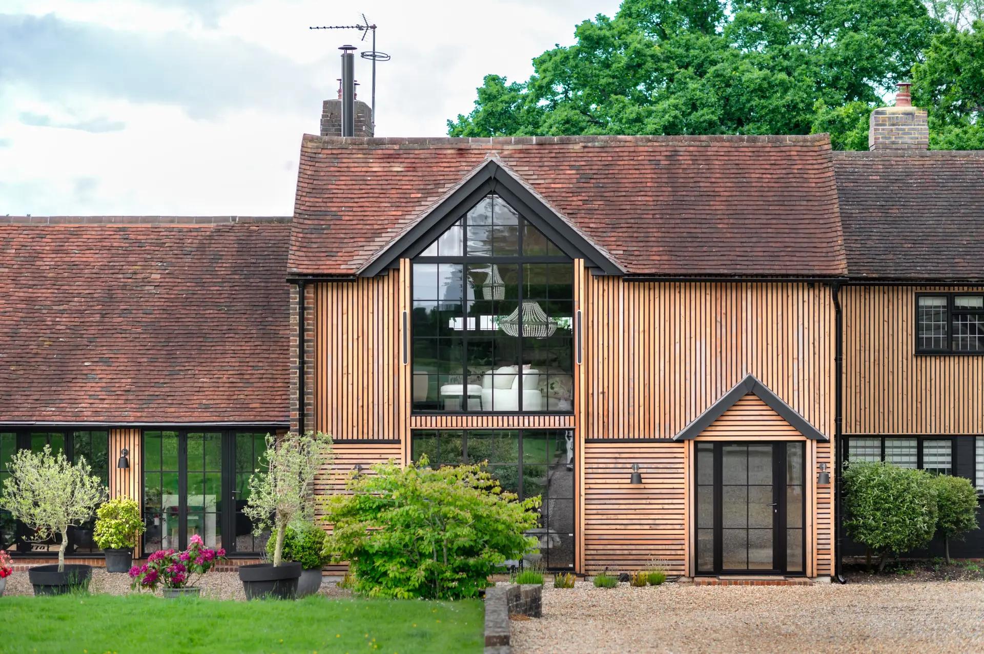 KP Glazing London A modern house with vertical wooden slats on the exterior, large glass windows, a gravel drive, and greenery including shrubs and potted plants in the front garden.