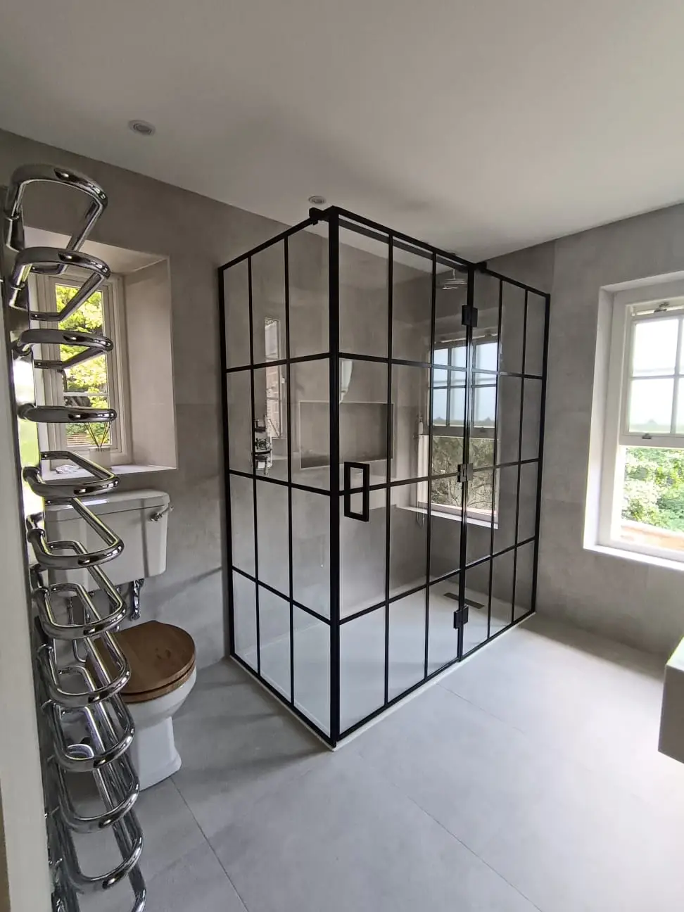 KP Glazing London A modern bathroom with a black-framed glass shower enclosure, a white toilet with a wooden seat, a chrome towel rail, light grey walls and floor, and a window letting in natural light.