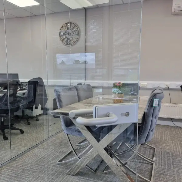 KP Glazing London A modern office meeting room with a glass door, grey chairs around a marble table, a wall clock, and computers visible through the glass wall. A small plant sits on the table.