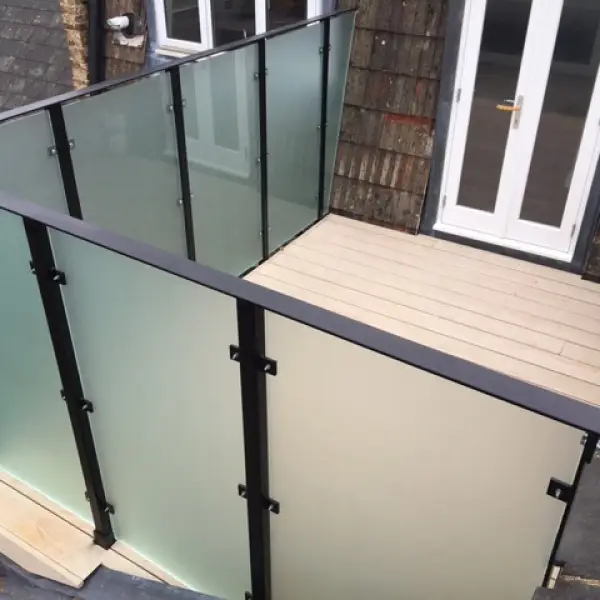 KP Glazing London A small balcony with frosted glass panel railings, black metal frames, and light-coloured wooden decking, attached to a building with white double doors and sloped tile cladding.