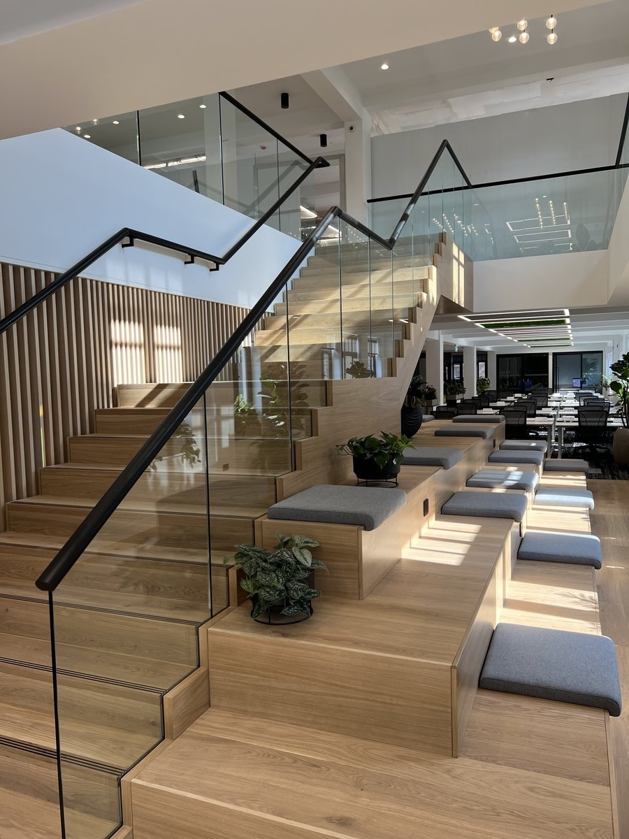 KP Glazing London Modern open office space with wooden tiered seating, grey seat cushions, glass railings, plants, and a staircase leading to an upper floor with glass walls and desks visible in the background. Natural light fills the area.