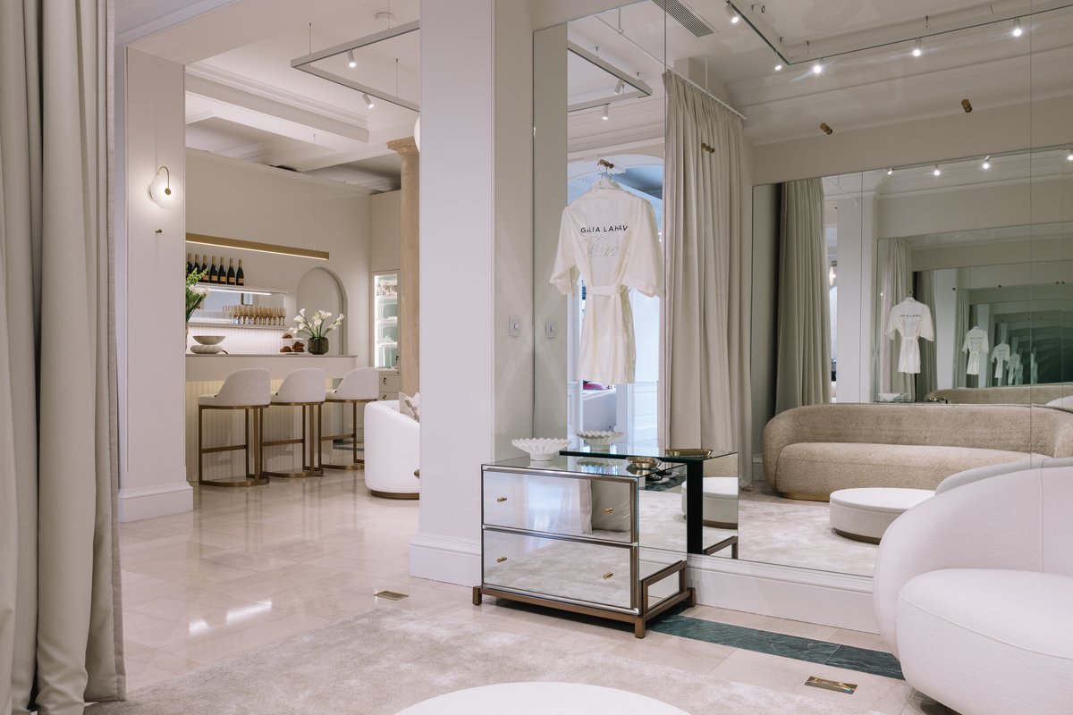 KP Glazing London A modern, elegant dressing room with a mirrored wall, beige sofa, glass drawers, and a white dressing gown hanging. In the background, there is a small bar area with stools, bottles, and soft lighting.