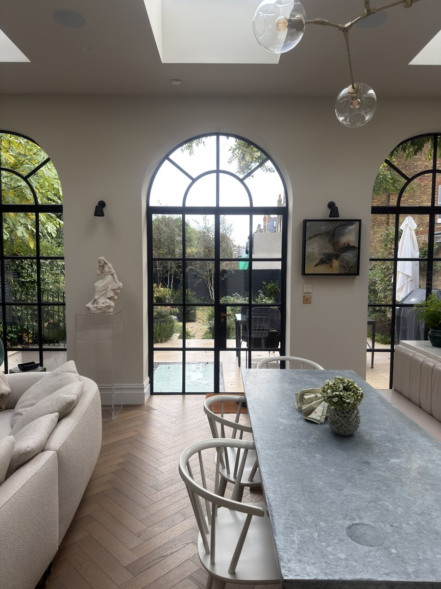 KP Glazing London A bright modern living and dining area with arched black-framed glass doors opening to a garden, herringbone wood flooring, light chairs around a grey stone table, a sofa, and decorative plants.