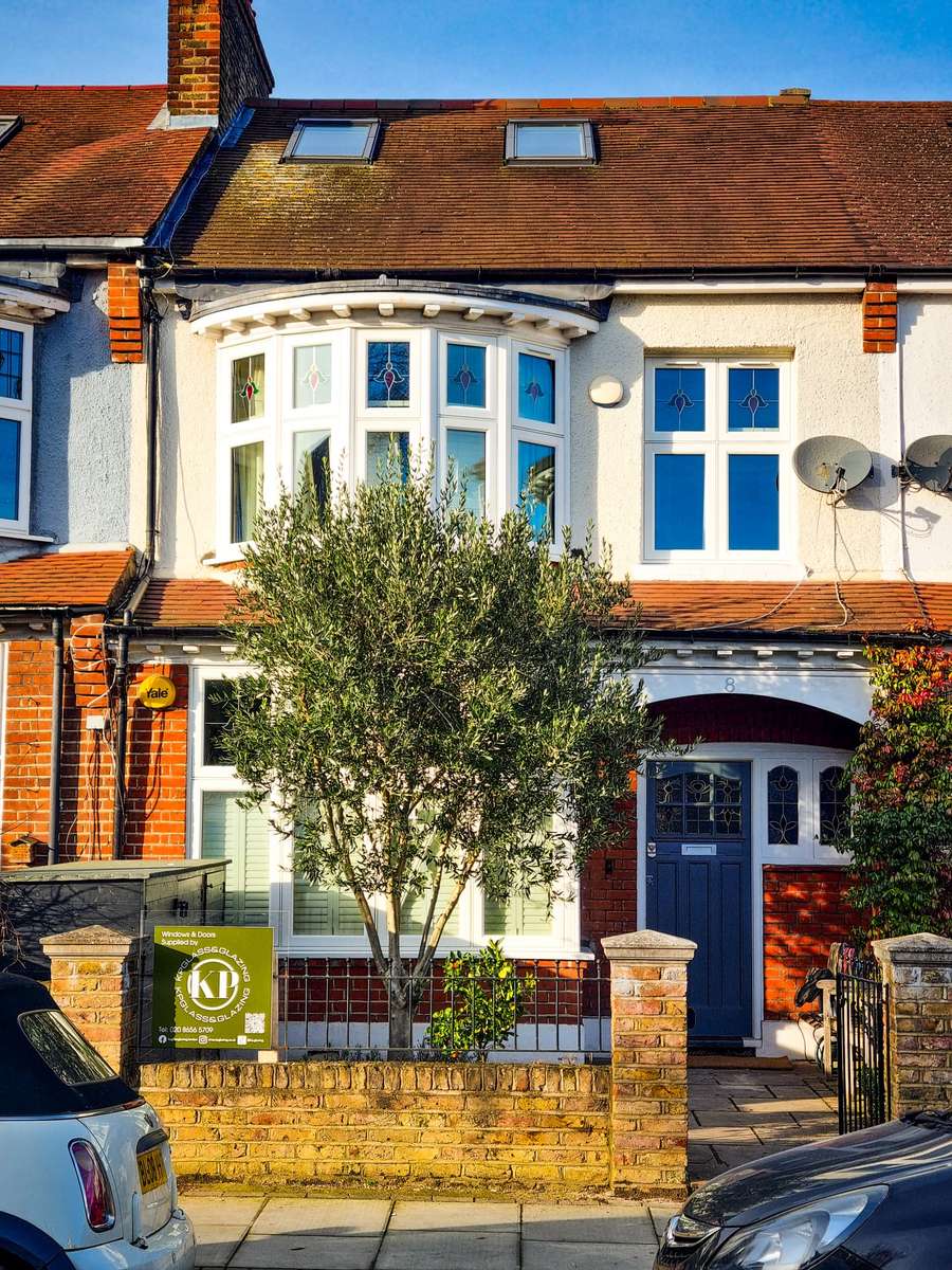KP Glazing London A brick terraced house with a bay window, blue door, two satellite dishes, a small front garden with a leafy tree, and parked cars on the street. A green security sign is visible on the front fence.