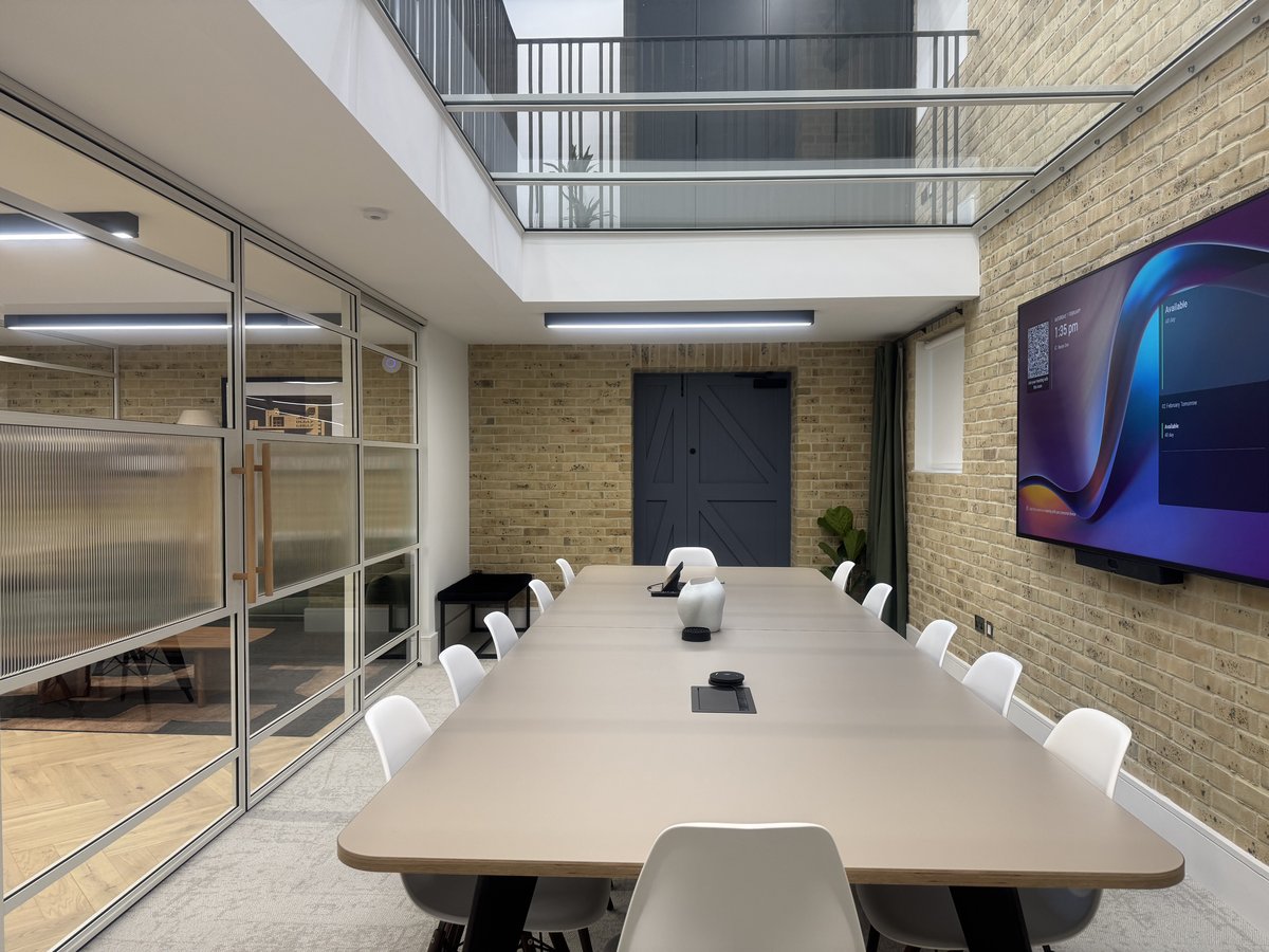 KP Glazing London A modern conference room with a long table, white chairs, a large wall-mounted screen, glass walls, brick walls, and a skylight letting in natural light. The room appears tidy and well-lit.
