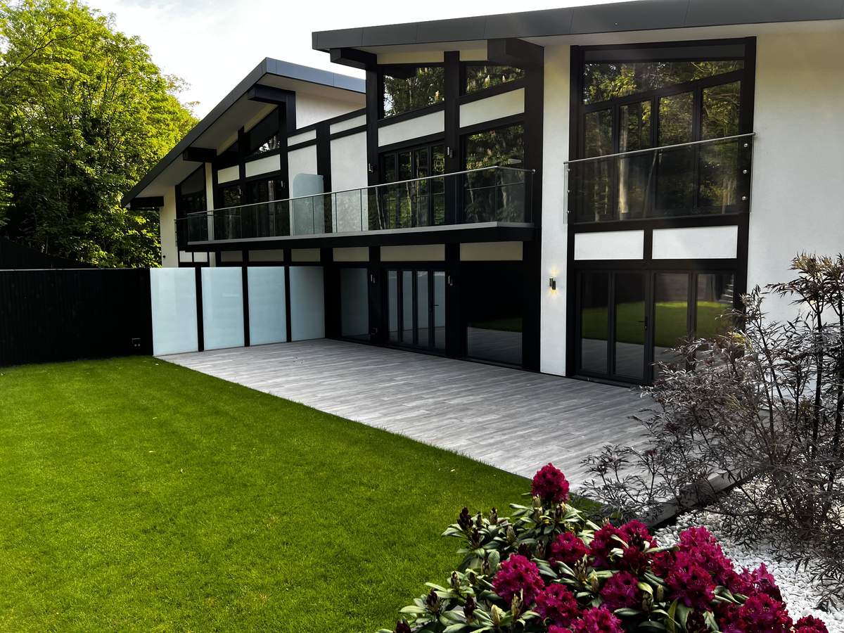 KP Glazing London Modern house with large windows and a glass balcony, featuring a spacious patio, green lawn, and blooming red flowers in the foreground. Lush trees and plants surround the contemporary building.