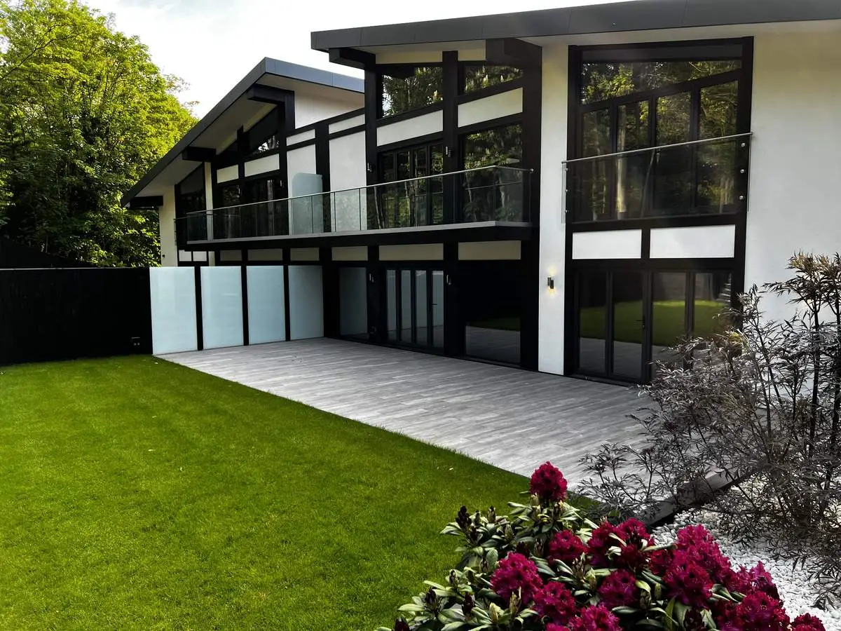 KP Glazing London Modern house with large windows and a glass balcony, featuring a spacious patio, green lawn, and blooming red flowers in the foreground. Lush trees and plants surround the contemporary building.
