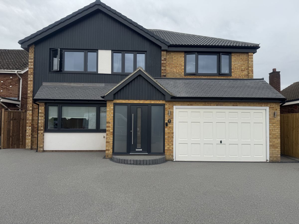 KP Glazing London Modern two-storey house with light brown brick and dark grey panelling, featuring large windows, a double front door with sidelights, and a white double garage door, all set on a wide, paved drive.