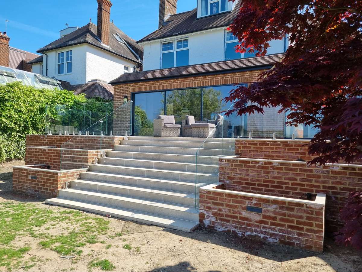 KP Glazing London A modern brick patio with wide stone steps, glass balustrades, and outdoor seating leads to a large house with big windows. A red-leaved tree is on the right side of the image.