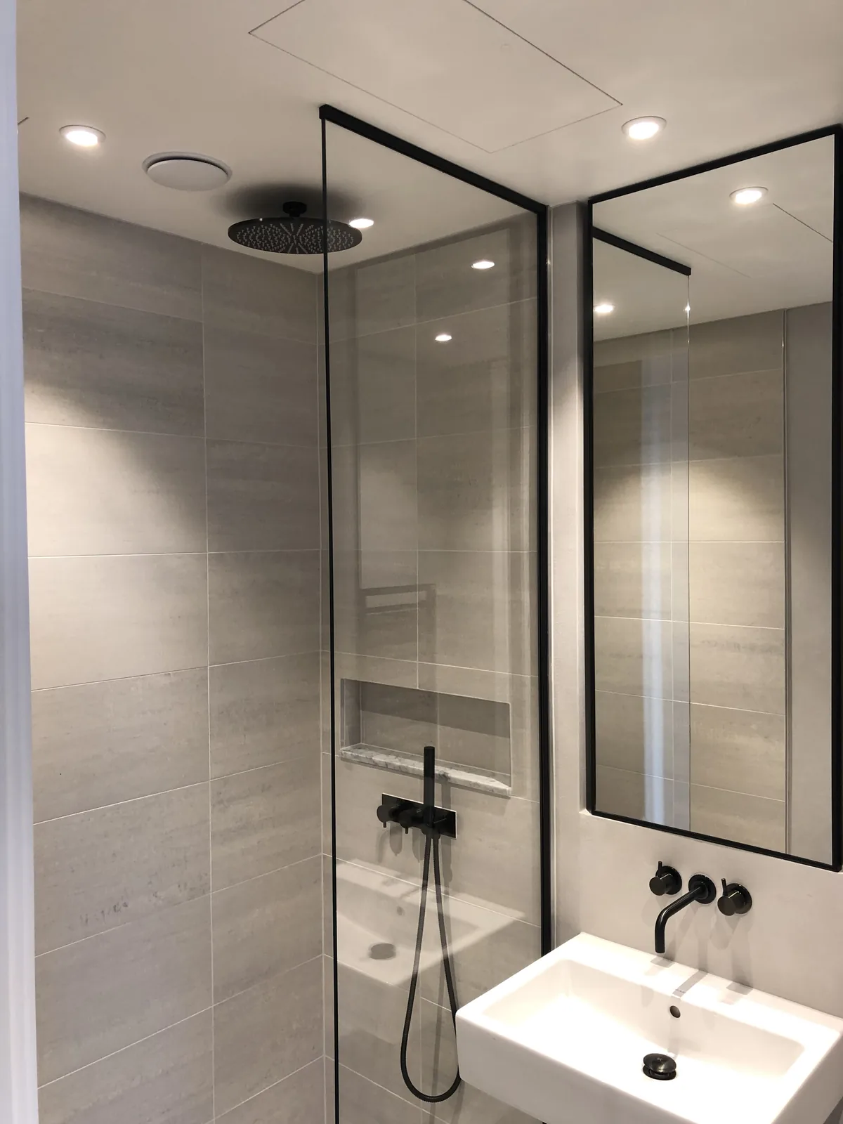 KP Glazing London Modern bathroom with light grey tiles, a walk-in glass shower, black fittings, a large mirror, and a white rectangular basin. Recessed ceiling lights provide illumination.