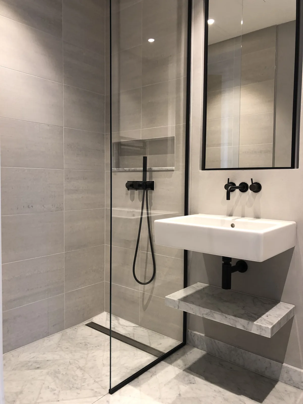 KP Glazing London Modern bathroom with light grey tiles, glass shower enclosure, black shower fittings, a white wall-mounted basin, and two marble shelves below the basin. Large mirror above the basin reflects the room.