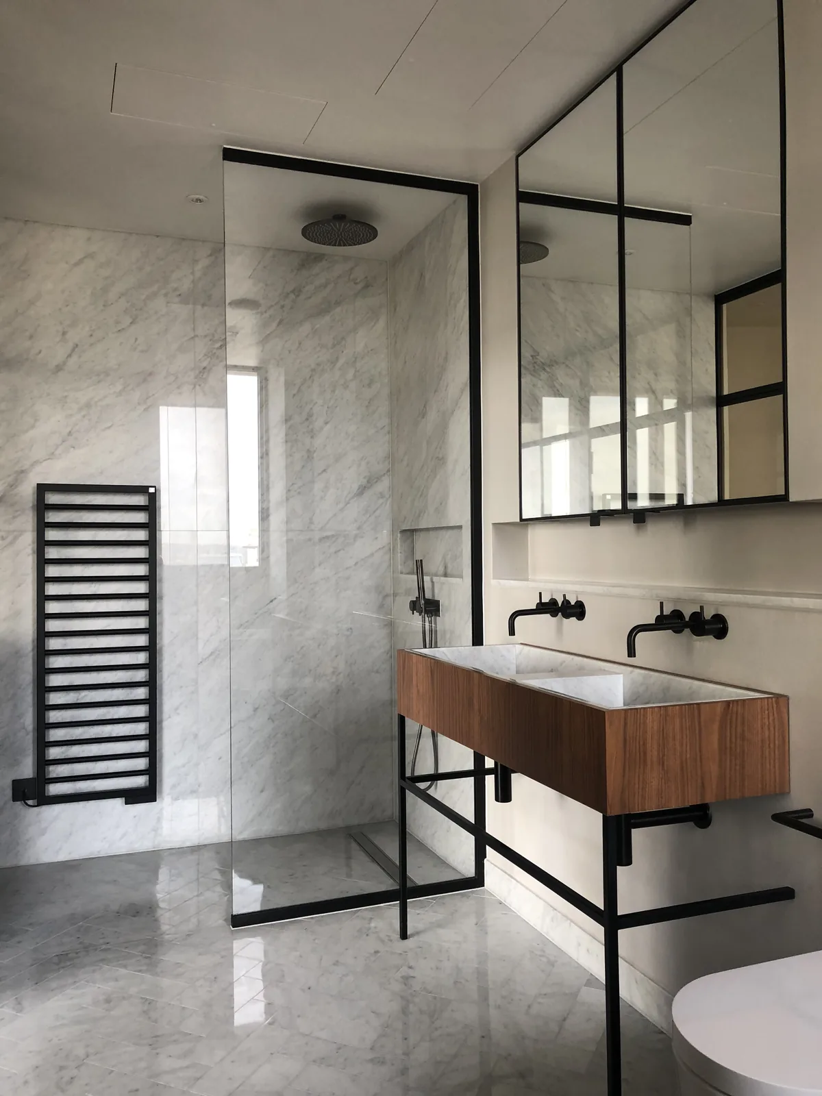 KP Glazing London Modern bathroom with marble walls and floor, glass walk-in shower, black fittings, twin basins with wooden vanity unit, large mirrored cupboards, black towel rail, and ceiling-mounted showerhead. Minimalist, clean design.