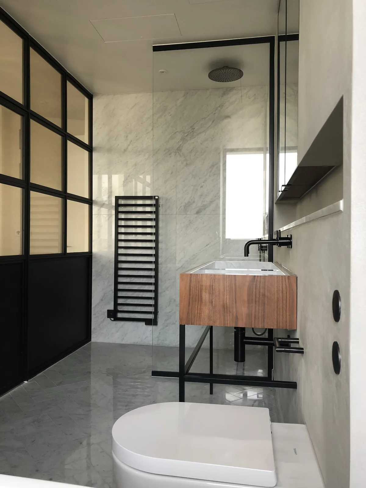 KP Glazing London Modern bathroom with marble walls and floor, glass shower enclosure, black towel rail, wooden floating vanity with basin, wall-mounted tap, and a white toilet in the foreground. Natural light enters through a window.