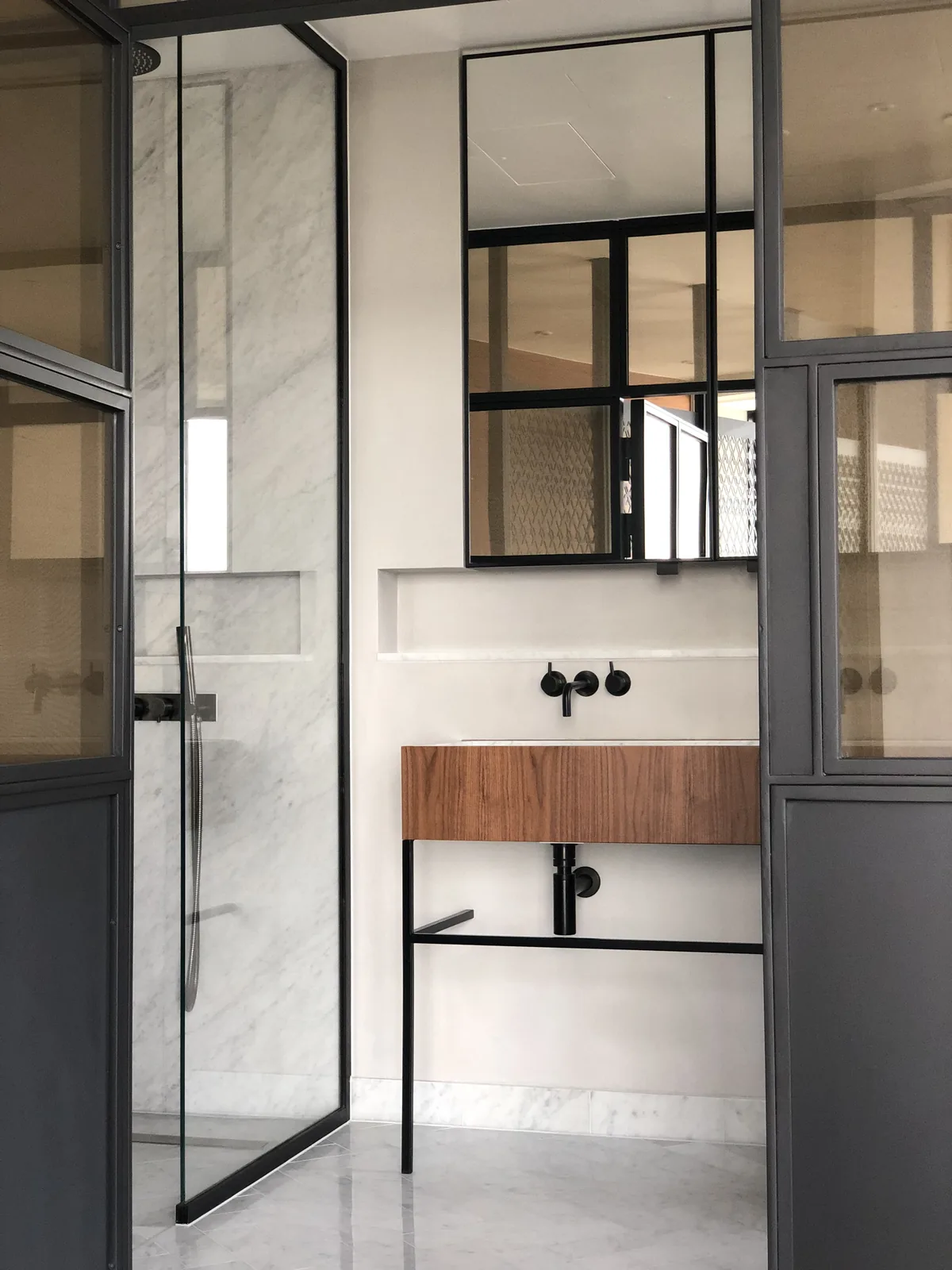KP Glazing London Modern bathroom with glass shower, marble walls and floor, wooden vanity with black fittings, large mirror, and black metal frame accents. Minimalist and clean design.