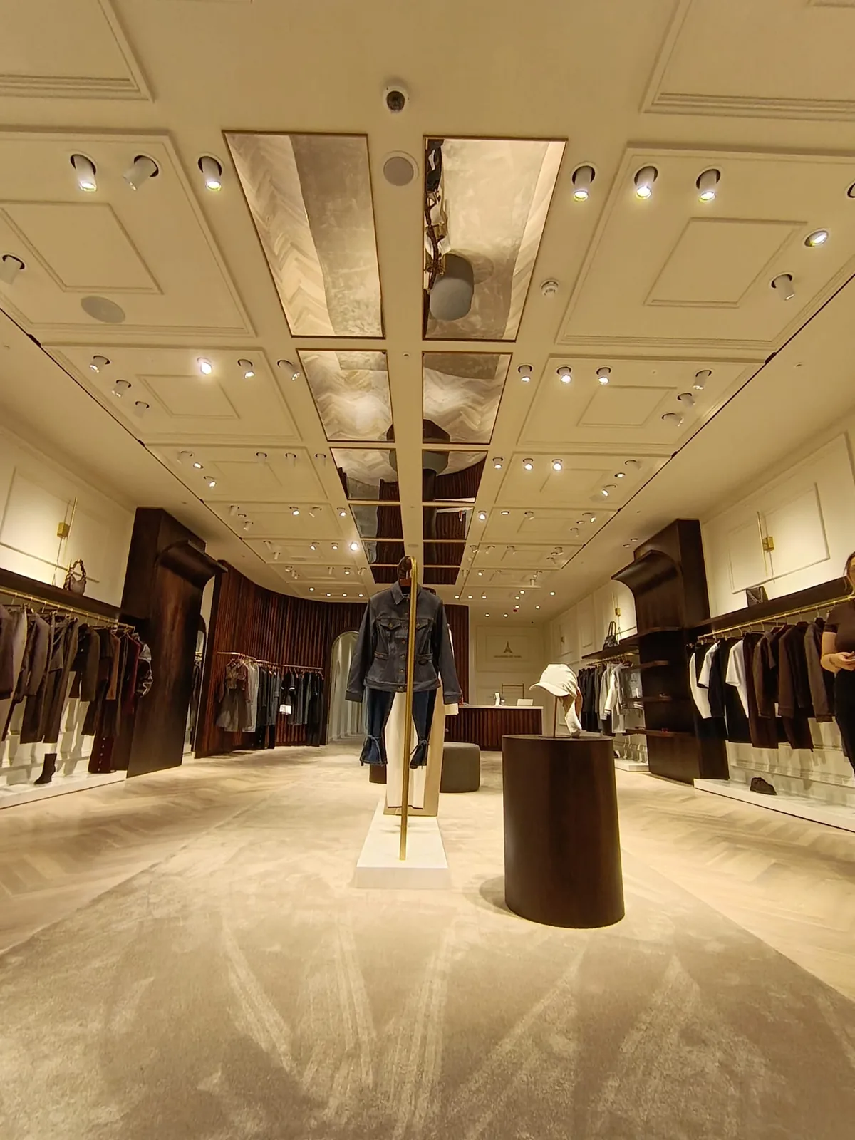KP Glazing London A luxury clothing shop interior with rails of clothes on both sides, a mannequin in the centre, plush beige carpet, spotlights, mirrors on the ceiling, and elegant dark wood accents.