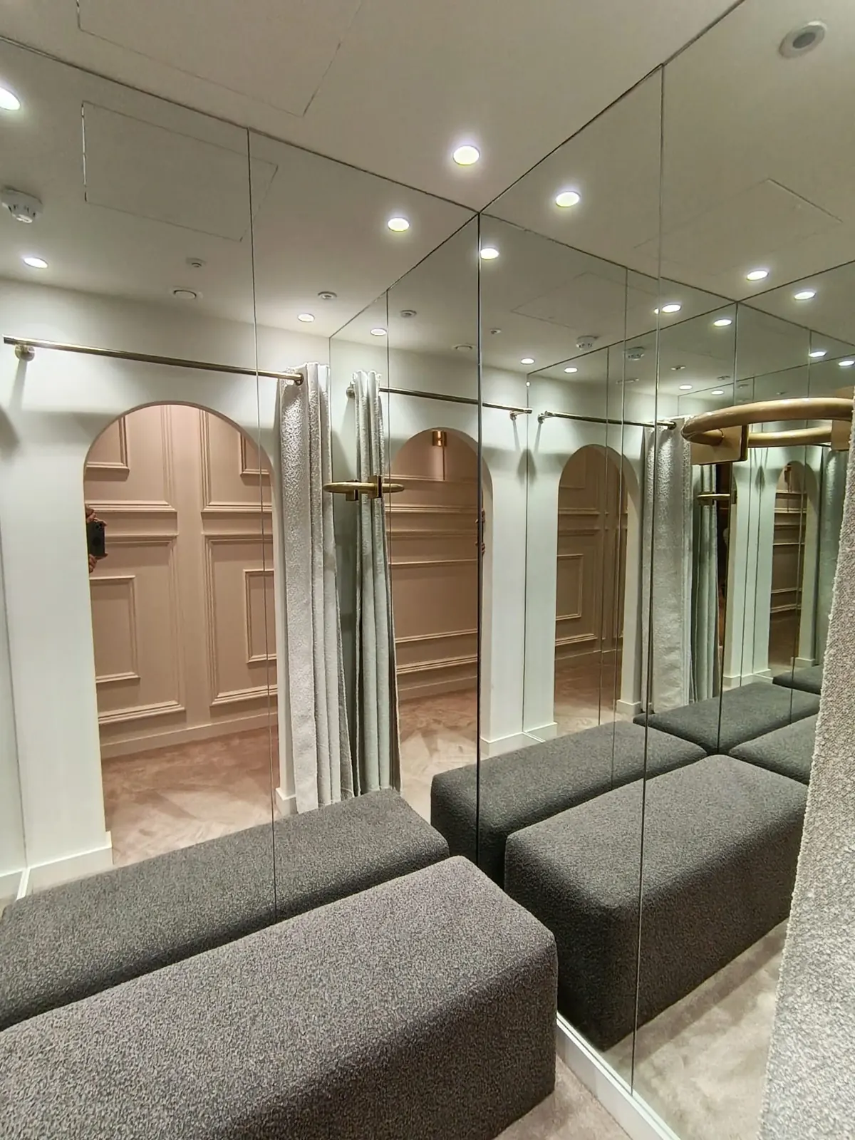 KP Glazing London A changing room with mirrored walls, grey benches, beige curtains, and arched panelling, creating the illusion of a larger, elegant space with soft lighting.