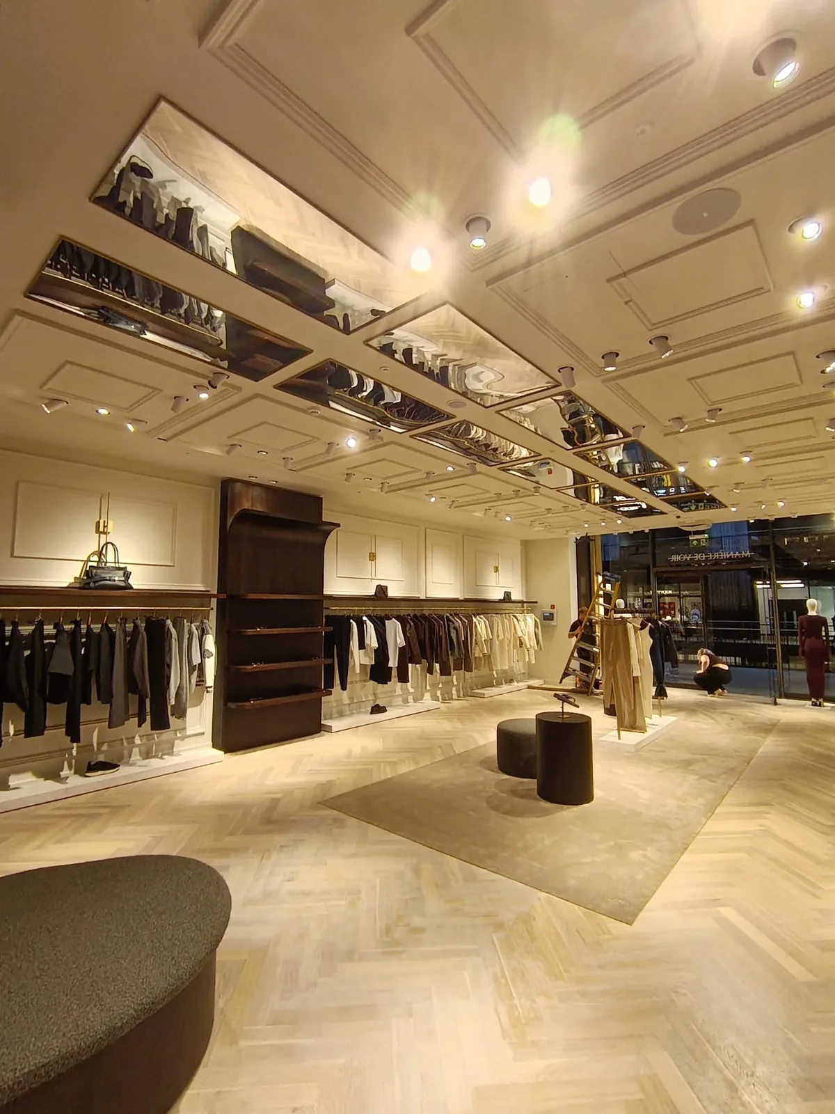 KP Glazing London A modern clothes shop with beige walls, hanging clothes, shelves, a soft bench, and mirrors on the ceiling. Bright lights illuminate the spacious, elegant interior.
