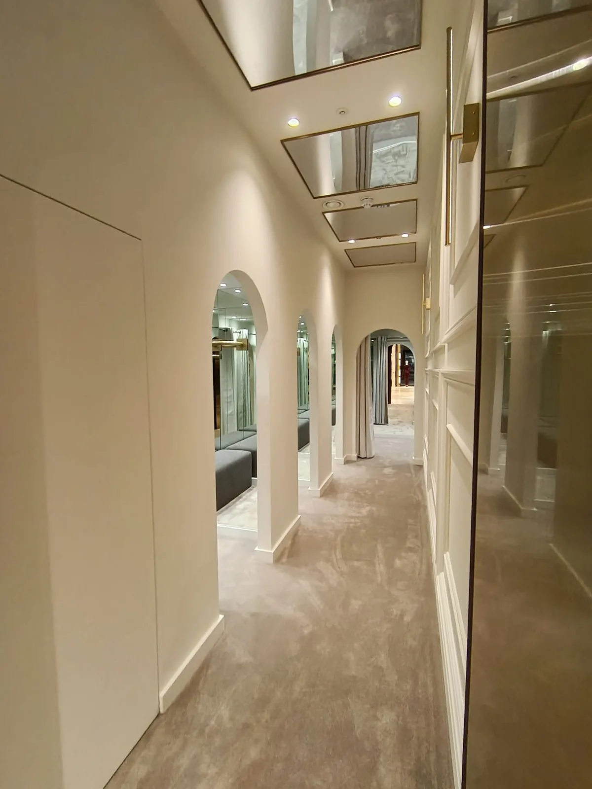 KP Glazing London A luxurious corridor with arched doorways, beige carpet, and mirrored ceiling panels. The space is well-lit with spotlights and features modern décor in neutral tones.