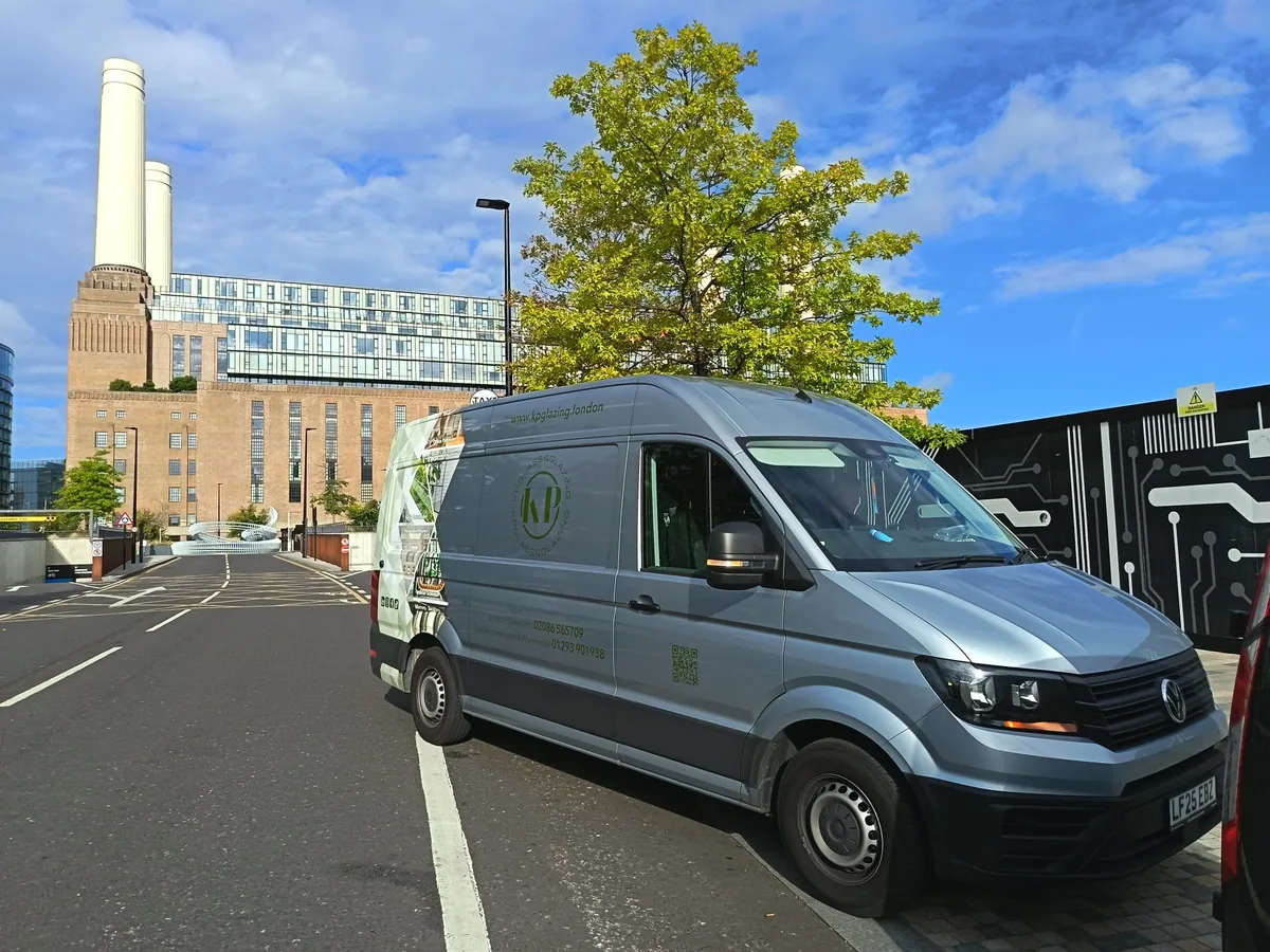 KP Glazing London A grey van is parked on a city street near a tall building with chimneys, a tree, and a blue sky with clouds in the background.